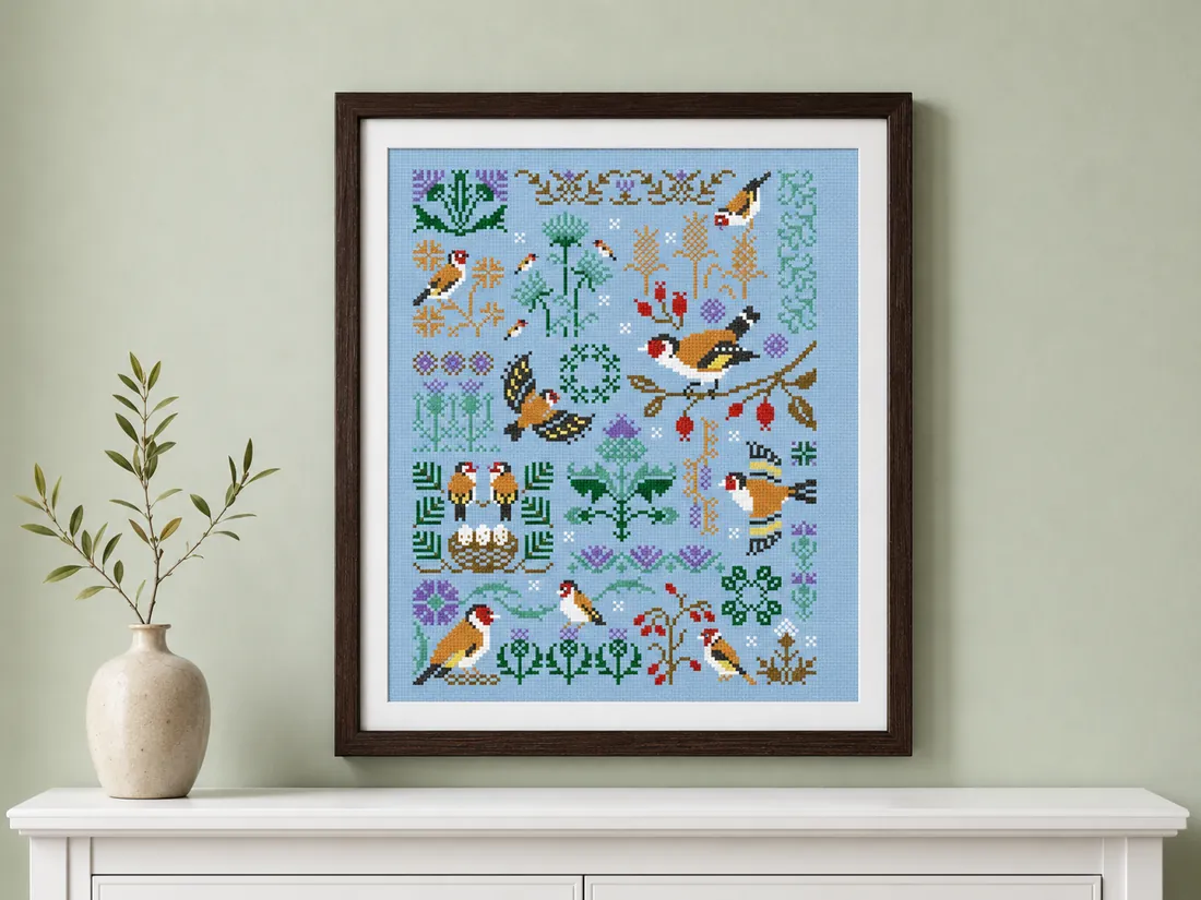 The finished Bird's Home sampler CC-K10-02 framed in dark brown wood on a pale sage wall above a white console, goldfinches around a nest of eggs on sky-blue Aida